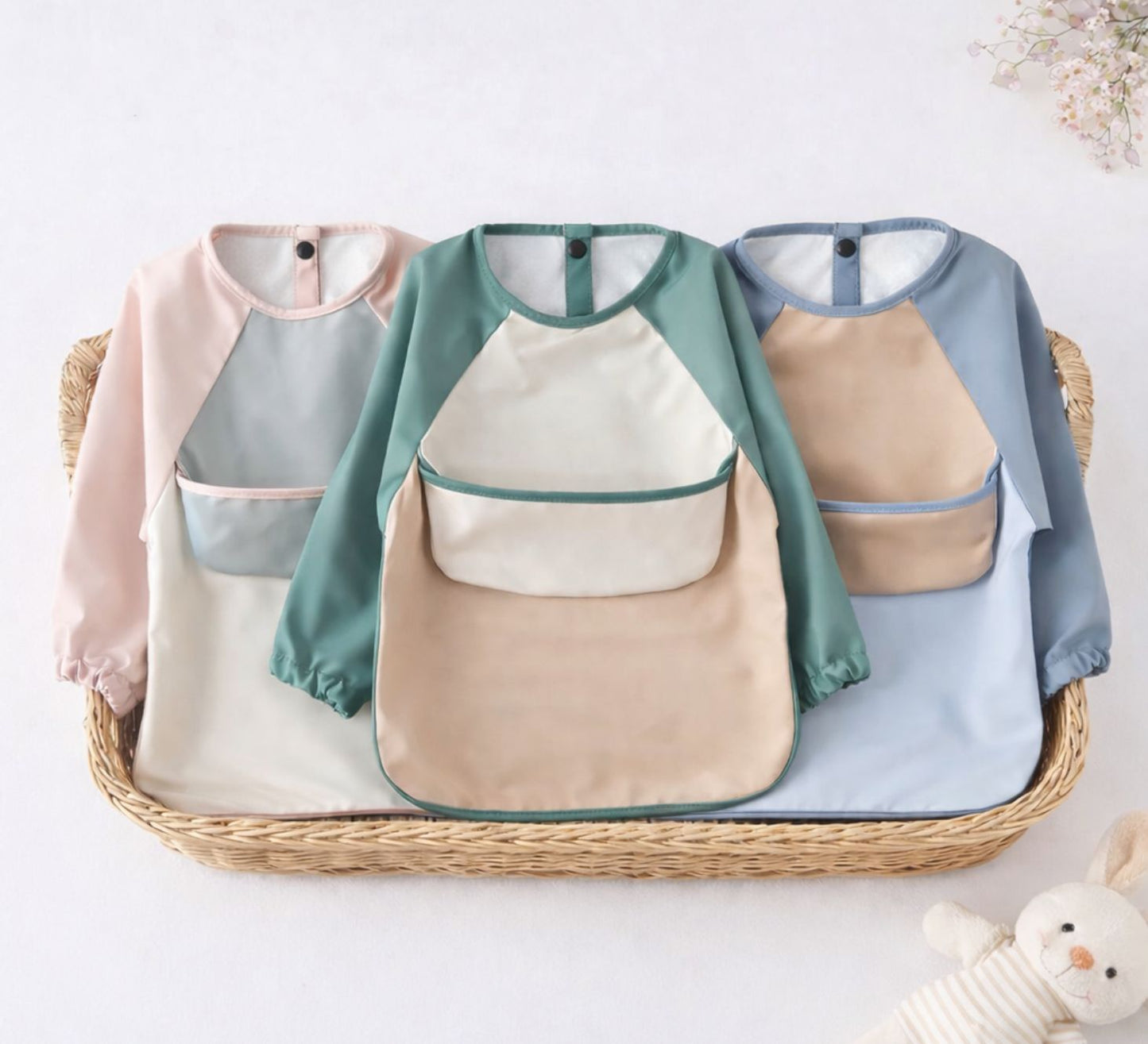 Set of waterproof baby bibs in pink, green and blue arranged in a basket on a light background