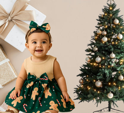 Baby in a green dress with gingerbread men pattern next to a decorated Christmas tree.