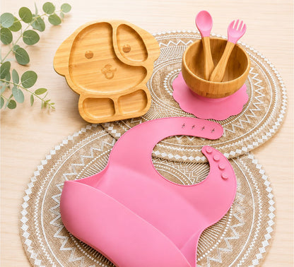 Organic bamboo dog-shaped baby feeding set with eco-friendly silicone bib, suction bowl, and wooden spoon and fork in pink colour.