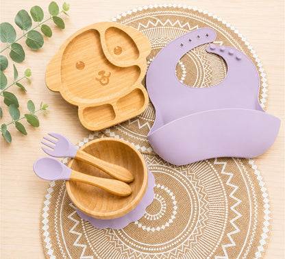 Organic bamboo dog-shaped baby feeding set with eco-friendly silicone bib, suction bowl, and wooden spoon and fork in purple colour.