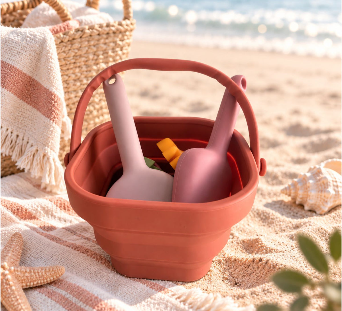 A terracotta-colored sand bucket with a handle, filled with pink and beige plastic beach toys, set on a sandy beach next to a woven basket and striped blanket.