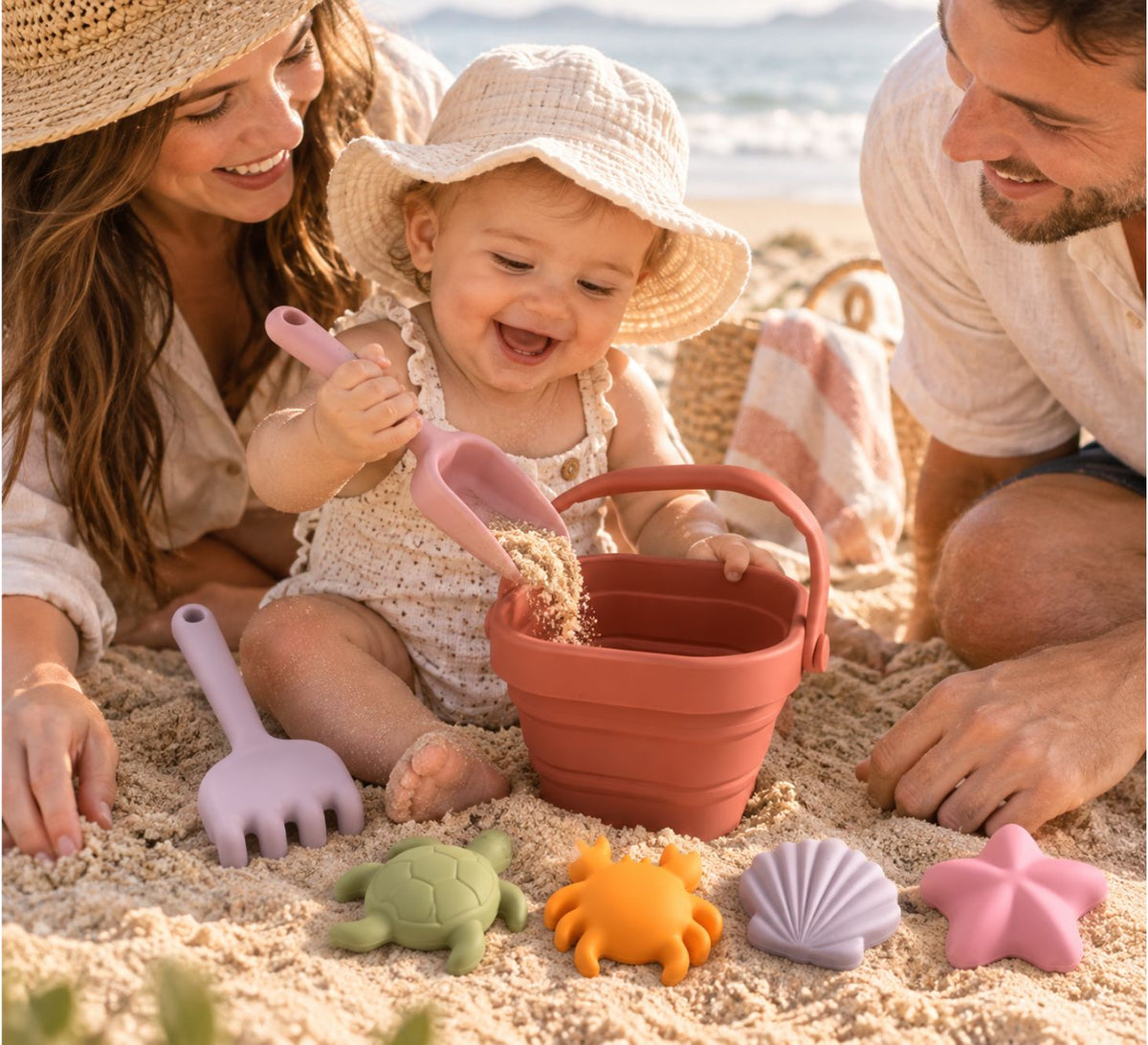 A baby sits on a sandy beach playing with a terracotta bucket, lavender rake, and colorful sand toys, surrounded by a smiling mother and father.