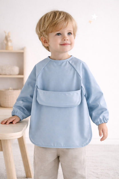 Child wearing a light blue apron standing in a room with a white wall and wooden furniture.